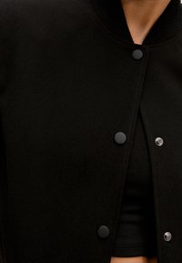 Schwarze Bomberjacke mit geripptem Kragen, Druckknöpfen und glatter Stoffoberfläche. Darunter ist ein schwarzes, eng anliegendes Oberteil sichtbar.