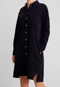 Abito camicia in corduroy blu navy con colletto, bottoni frontali, due tasche sul petto e spacchi laterali. Maniche lunghe, con bottoni sui polsini.