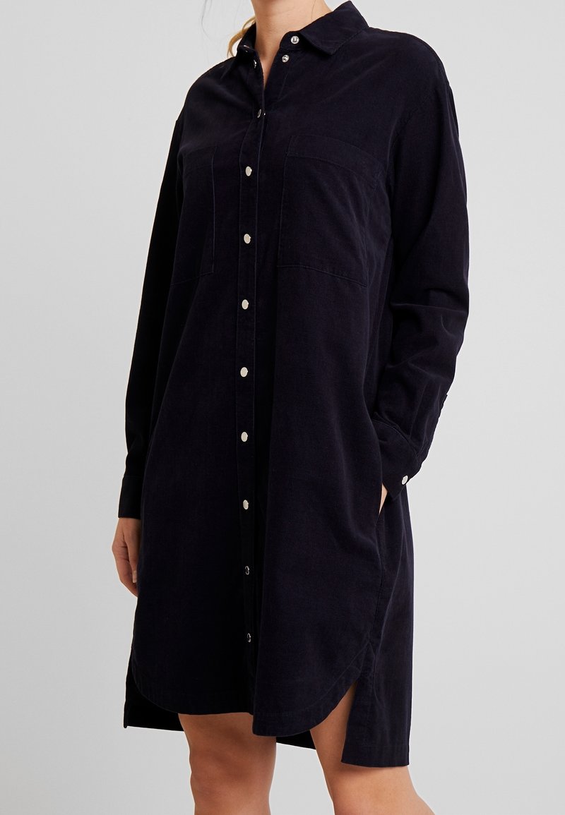 Abito camicia in corduroy blu navy con colletto, bottoni frontali, due tasche sul petto e spacchi laterali. Maniche lunghe, con bottoni sui polsini.