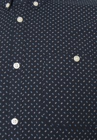 Námořnická modrá košile s knoflíky vyrobená z látky s jemným bílým a modrým vzorem. Obsahuje kapsu a knoflíky připomínající perly.