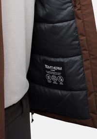 Giacca isolante marrone con interno nero trapuntato. Presenta un'etichetta con istruzioni per la cura e icone per calore, traspirabilità e asciugatura.