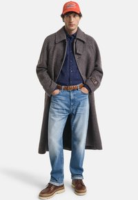 Bruine houndstooth jas, marineblauwe overhemd, lichtblauwe jeans, bruine schoenen met crèmekleurige zolen en een rode pet met witte tekst.