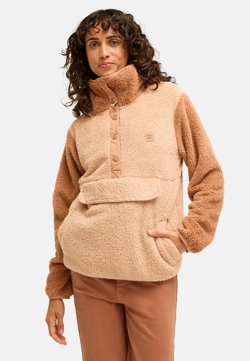 Femme aux cheveux bouclés portant un pull polaire bicolore beige avec col boutonné et poche avant, associé à un pantalon marron.