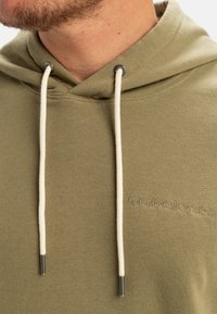 Olive green hoodie in soft fabric, featuring a front kangaroo pocket, cream drawstrings, and embroidered logo in matching color.