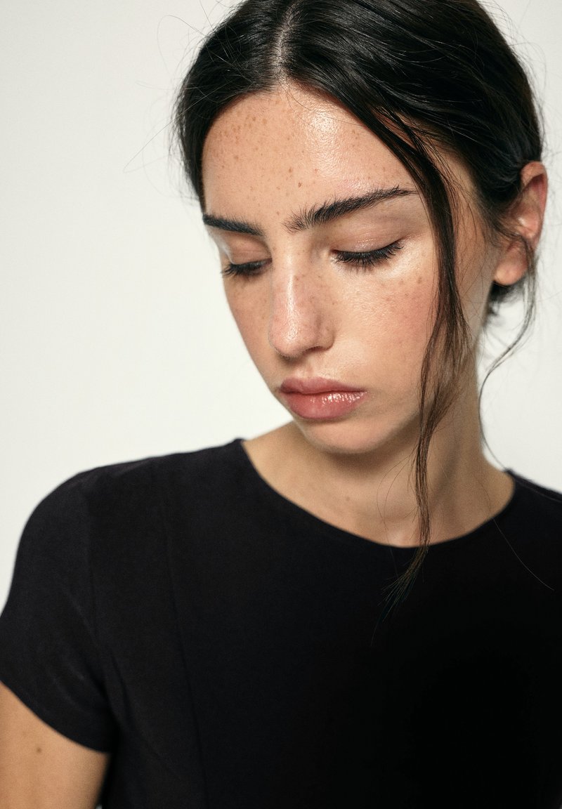 Jeune femme aux cheveux foncés attachés en arrière, portant une chemise noire, regardant vers le bas avec une expression neutre sur un fond uni.
