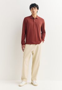 Polo de manga larga color óxido con cuello y tapeta de tres botones, combinado con pantalones beige claro y zapatillas blancas.