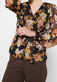 Blumenbluse mit schwarzem Hintergrund, auf dem gelbe, rosa und weiße Blumen abgebildet sind, langen transparenten Ärmeln und einem Bindeband in der Taille.