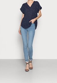 Blouse bleu marine à pois blancs, décolleté en V, manches courtes retroussées ; associée à un jean skinny bleu clair et des sandales à talons noires.