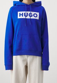 Niebieska bluza z kapturem ze ściągaczem, duże białe logo "HUGO" na przodzie, kieszeń kangurka, ściągacze na mankietach i u dołu, noszona na jasnych spodniach.
