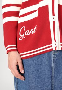 Rød cardigan med hvite striper, med knappelukking og en lomme. "Gant" er brodert i hvitt foran. Mykt strikket materiale.