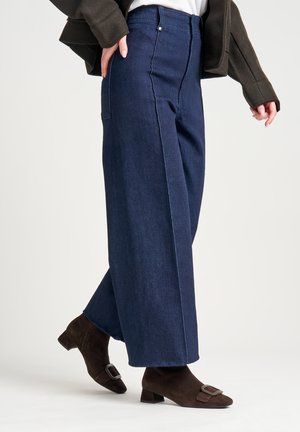 Pantalones de mezclilla oscuros de pierna ancha con detalles de costura visibles y dos bolsillos laterales, combinados con botas de ante marrones hasta el tobillo con una hebilla.