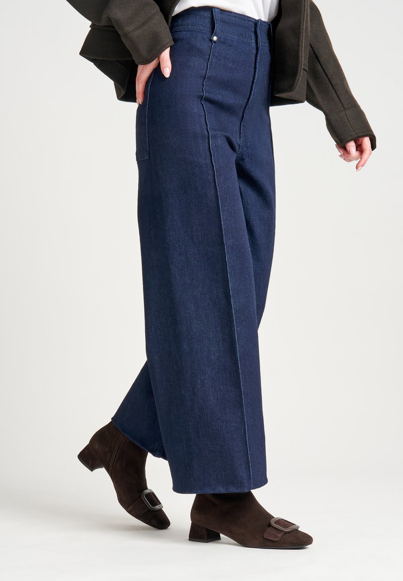 Pantalones de mezclilla oscuros de pierna ancha con detalles de costura visibles y dos bolsillos laterales, combinados con botas de ante marrones hasta el tobillo con una hebilla.