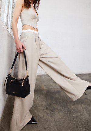 Kobieta w beżowych spodniach o szerokich nogawkach i skróconej kamizelce, trzymająca czarną torebkę, stojąca na betonowej podłodze na tle białej ściany.