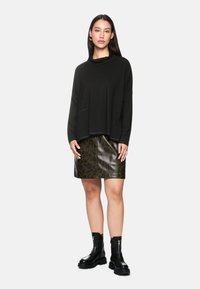 Frau in einem lockeren, schwarzen Langarmoberteil, einem Leopardenmuster-Lederminirock, schwarzen klobigen Stiefeln und goldenen Ohrringen, die vor einem weißen Hintergrund steht.