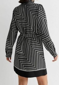 Donna che indossa un abito a maniche lunghe a strisce geometriche bianche e nere, con vita cintrata e orlo nero, rivolta lontano dalla macchina fotografica.