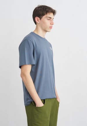 Jonge man in een blauw T-shirt met korte mouwen en olijfgroene broek staat zijwaarts met handen in de zakken tegen een effen witte achtergrond.