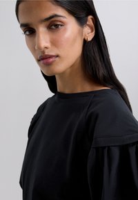 Ung kvinna med långt mörkt hår, iklädd en svart topp med pufsig ärm, som ser något åt sidan mot en enfärgad ljus bakgrund.