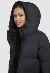 Sort puffjakke med en stor hætte, lavet af glat nylon. Har en lynlåslukning fortil, knapper og et blødt indre foer for varme.