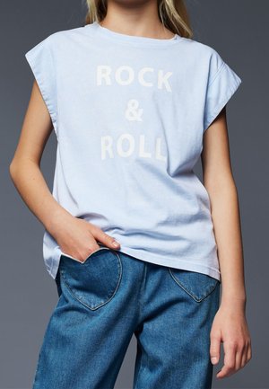 Kobieta ubrana w jasnoniebieską koszulkę bez rękawów z napisem "ROCK & ROLL" oraz niebieskie dżinsy z kieszeniami z przodu w kształcie serca, jedną ręką w kieszeni.