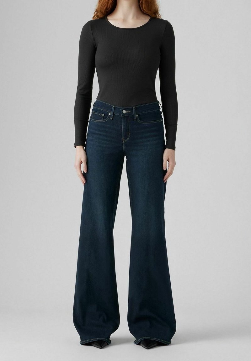 Femme portant un haut noir moulant à manches longues et un jean bleu foncé taille haute, jambes larges, debout devant un fond gris.