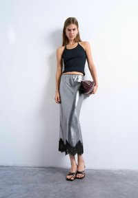 Femme portant un haut noir sans manches, une jupe midi argentée avec un ourlet en dentelle noire, des sandales noires, tenant une pochette bordeaux cloutée, debout contre un mur blanc.