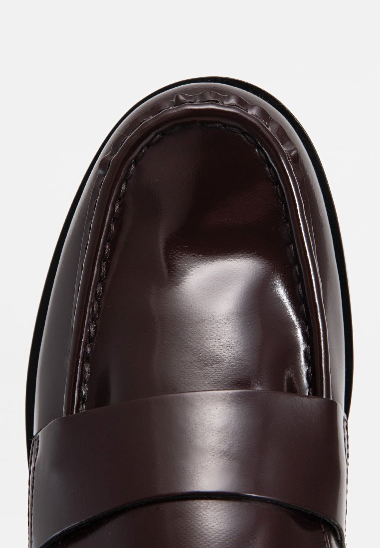 Brązowy, błyszczący skórzany mokasyn o gładkiej powierzchni, zaokrąglonym czubku i ozdobnej taśmie z przodu. Detale szwu wokół krawędzi.