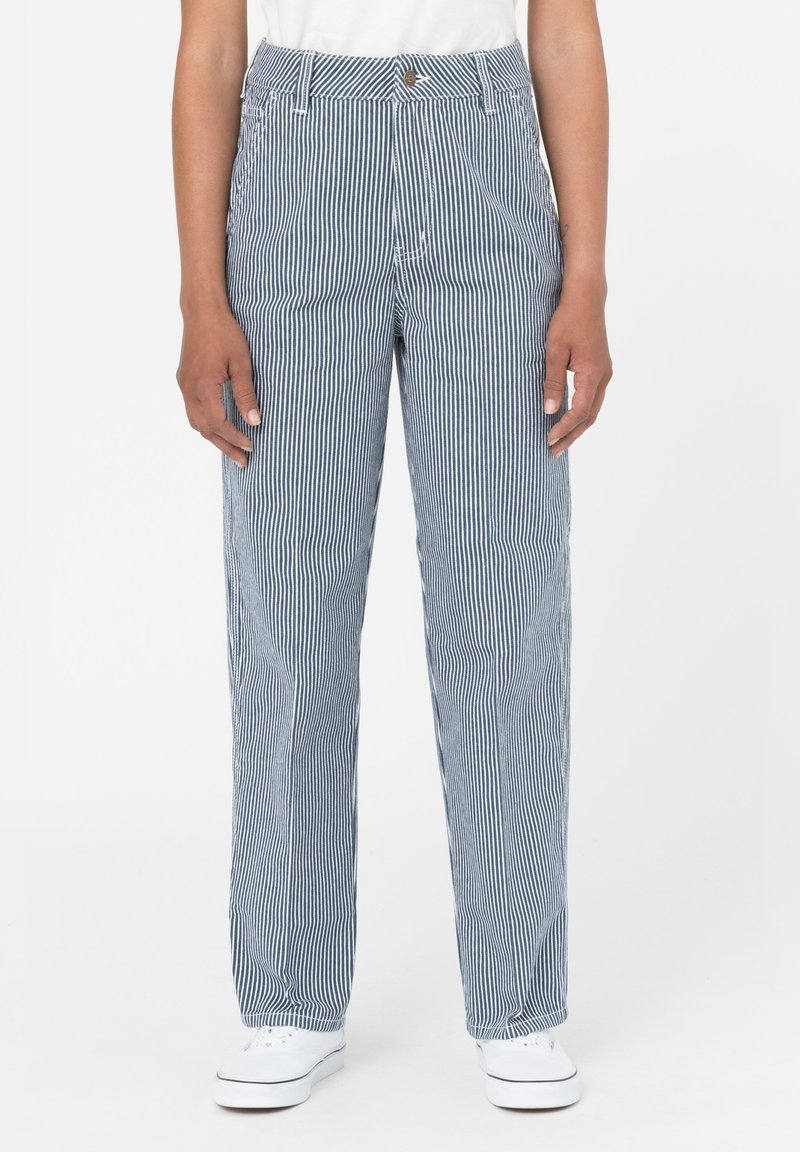Personne portant un pantalon taille haute, jambes droites, avec des fines rayures verticales bleues et blanches et des baskets blanches, debout sur un fond blanc.