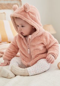 Hoodie rose clair et doux avec des oreilles d'ours, doté d'une fermeture éclair à l'avant. Porté avec un pantalon de pyjama blanc à fleurs et des chaussettes beiges en tricot.