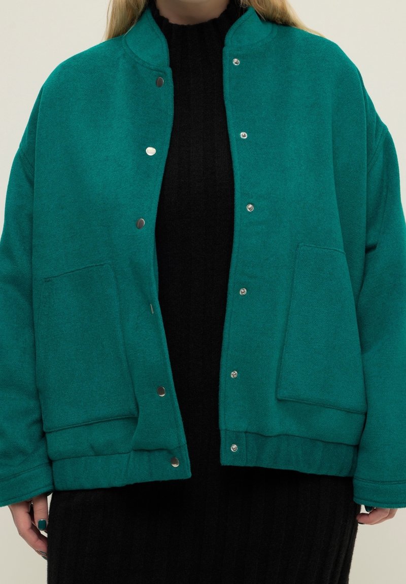 Veste en laine bleu sarcelle avec un col arrondi, des boutons pression, deux poches frontales et une coupe décontractée. Portée sur une robe noire côtelée.