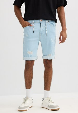 Hombre con pantalones cortos de mezclilla enrollados de color azul claro con cordón, camiseta negra, calcetines blancos y zapatillas blancas, de pie frente a un fondo blanco.