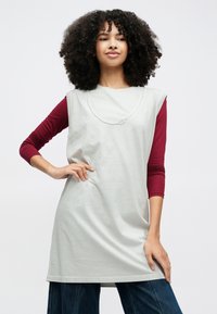 Sieviete ar cirtainiem matiem, valkā bezroku gaiši pelēku tuniku pār garām piedurknēm bordo tops un zilas džinsa bikses, pārliecinoši pozējot ar roku uz gurna.