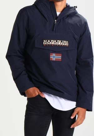 Mand iført navyblå Napapijri hættetrøje med frontlomme og norsk flagpatch, kombineret med sorte bukser og hvid skjorte.