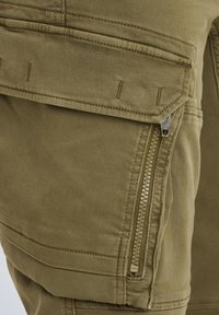 Maslinaste zelene cargo hlače s patentnim zatvaračem, šivenim detaljima i izdržljivim materijalom od pamuka. Dizajn uključuje teksturiranu površinu.