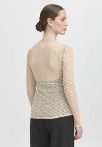 Spitzenoberteil in Nudefarbe, lange Ärmel, taillierte Passform, strukturiertes Blumenmuster, gewellter Saum, getragen mit schwarzen Hosen.