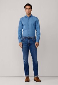 Lichtblauw overhemd met knopen en lange mouwen, gecombineerd met donkeblauwe spijkerbroek. Het geheel wordt compleet gemaakt met bruine schoenen, met een klassieke pasvorm.