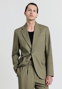 Giacca da suit verde oliva con rever a punta, due bottoni e taschino sul petto. Tessuto liscio e vestibilità su misura, abbinata a una camicia nera.