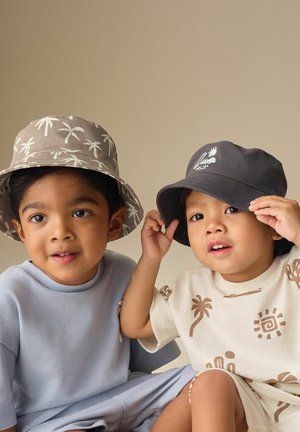 Kaksi nuorta lasta, jotka molemmat käyttävät lierihattuja; toinen on harmaa valkoisilla palmupuilla, toinen musta valkoisella logolla, istuvat ja katsovat eteenpäin.