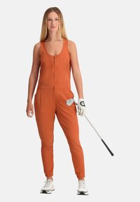 Tuta arancione senza maniche con zip frontale, polsini elasticizzati e tasche laterali, indossata con una mazza da golf e un guanto. Sneaker bianche completano il look.