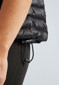 Schwarze Pufferjacke mit glatter, glänzender Textur, versehen mit einem Kordelzug und einem Etikett "JACKIT" am Saum, getragen über schwarzen Leggings.