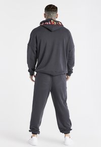 Felpa con cappuccio grigio scuro con interno del cappuccio a motivo, pantaloni della tuta abbinati e polsini a coste, presentata da dietro. Scarpe da ginnastica bianche visibili.