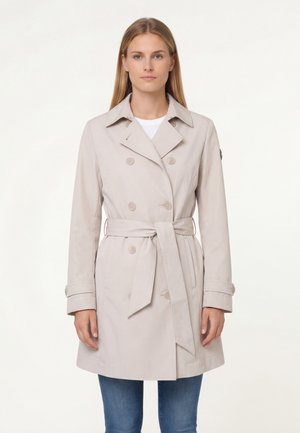 Donna che indossa un trench doppiopetto beige con cintura in vita e cinturini ai polsi, abbinato a jeans blu, in piedi davanti a uno sfondo neutro.