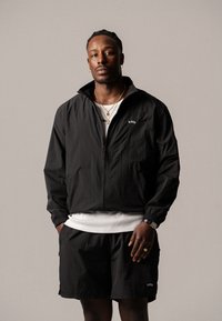 Giacca leggera nera con zip e polsini elasticizzati, abbinata a pantaloni corti coordinati. Entrambi presentano un logo discreto, indossati sopra una maglietta bianca.