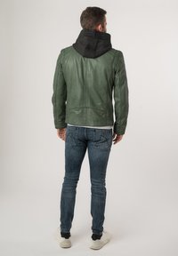 Giacca di pelle verde con cappuccio nero, design aderente e dettagli di cucitura. Abbinata a jeans blu e sneakers bianche.