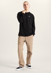 Maglietta nera a maniche lunghe con logo sul petto, abbinata a chinos color beige e scarpe da ginnastica nere. L'outfit presenta elementi di design semplici e casual.