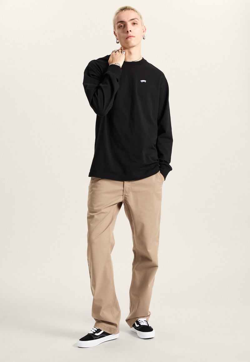 Maglietta nera a maniche lunghe con logo sul petto, abbinata a chinos color beige e scarpe da ginnastica nere. L'outfit presenta elementi di design semplici e casual.