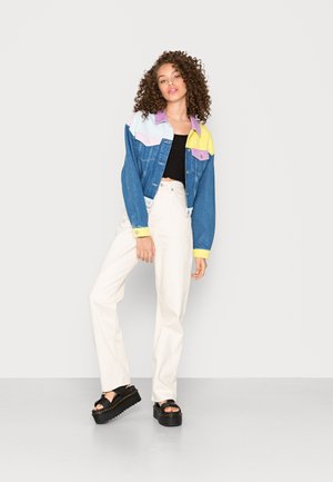 Jaqueta de ganga com colarinho em blocos de cores rosa, roxo e amarelo; combinada com um top cropped preto, calças largas em creme e sandálias de plataforma.