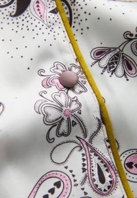 Bluza cu mâneci lungi, confecționată dintr-un material crem deschis, având un imprimeu floral și paisley în nuanțe de maro și roz, evidențiată printr-o margine și nasture galbene.