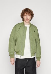 Giacca bomber verde con chiusura a zip, polsini a coste e logo ricamato sul petto, indossata sopra una t-shirt bianca.