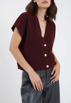 Donna che indossa un gilet senza maniche rosso scuro con bottoni e pantaloni in pelle neri, con le mani appoggiate sui fianchi su uno sfondo semplice.