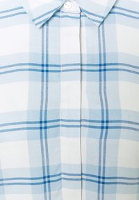 Camicia a quadro azzurro chiaro e bianco con colletto classico, pattina dei bottoni e texture di tessuto morbido con linee orizzontali e verticali ben definite.